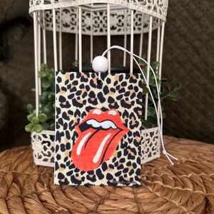 May include: Car air freshener with a red and white tongue graphic on a leopard print background. The air freshener is hanging from a white string with a white bead.