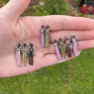 May include: Three sets of silver pins depicting three figures standing together, each wearing a different coloured dress. The dresses are pink, green, and brown. The figures are holding hands and have a heart on their chest.