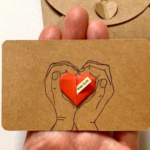 Könnte beinhalten: Eine braune Karte mit einer handgezeichneten Umrisszeichnung von Händen, die ein rotes Origami-Herz halten. Das Herz hat ein kleines weißes Etikett mit dem Text "Alles Gute". Ein passender Umschlag ist im Hintergrund.