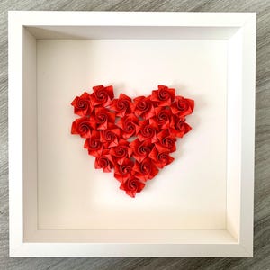 Könnte beinhalten: Ein gerahmtes Kunstwerk mit einem Herzen aus kleinen, roten Papierrosen. Das Herz befindet sich auf einem weißen Hintergrund in einem weißen quadratischen Rahmen. Die Rosen sind leuchtend rot und bilden einen Kontrast.