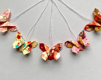 May include: Five origami butterflies hanging from white strings against a white background. The butterflies are made from patterned paper in shades of pink, red, yellow, and brown.