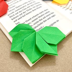 Könnte beinhalten: Ein grünes Origami-Lesezeichen in Form eines vierblättrigen Kleeblatts liegt auf der Ecke eines geöffneten Buches. Das Buch ist auf einer Seite mit deutschem Text über einen Hund geöffnet.