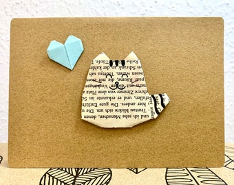 May include: A handmade card with a light blue origami heart and a white origami cat made from book pages. The card is on a brown background with a black and white pattern.