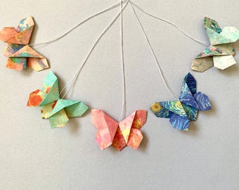 May include: A collection of six origami butterflies in various colors and patterns, suspended by white strings. The butterflies feature shades of orange, pink, blue, and green, with some showcasing artistic designs. The butterflies are arranged against a light gray background.