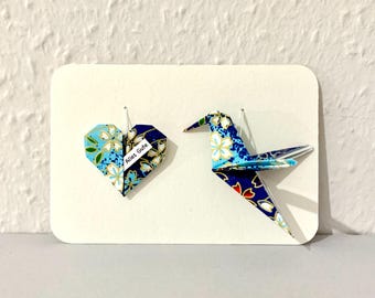 May include: A white card features a blue and white origami heart and crane. The heart has a floral pattern and a small label with text. The crane is also blue and white with a floral design. Both are attached to the card with pins.