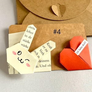 Puede incluir: Un conejo de origami hecho de páginas de libro color crema con texto en tinta negra y mejillas rosadas, colocado junto a un corazón de origami rojo con el texto Ich liebe dich en una tarjeta marrón etiquetada #4, con un sobre marrón de fondo.