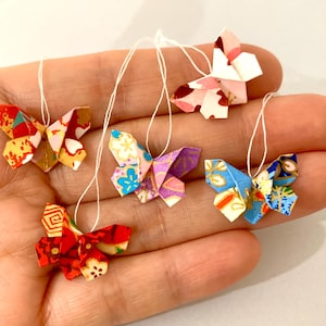 Könnte beinhalten: Sechs bunte Origami-Schmetterlinge, jeweils an einem weißen Faden aufgehängt, werden in einer Hand präsentiert. Die Schmetterlinge weisen komplizierte Faltmuster und eine Vielzahl von Farben auf, die die Kunst des Papierfaltens zeigen.