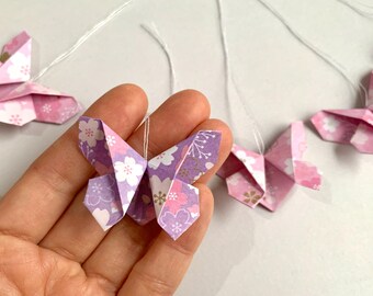 May include: A garland of five origami butterflies with a floral pattern in shades of pink and purple. Each butterfly is attached to a white string.