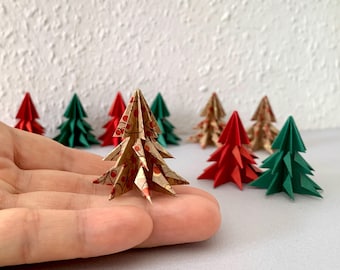 Könnte beinhalten: Eine Sammlung von Miniatur-Origami-Weihnachtsbäumen in verschiedenen Farben und Mustern. Ein Baum besteht aus beigem Papier mit einem roten Beeren- und Rankenmuster, während andere einfarbig rot, einfarbig grün oder mit goldenem Blumenmuster sind.