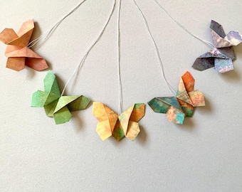 May include: Origami butterfly decorations in various colors, including green, orange, and purple, suspended by white strings. The paper butterflies are folded into detailed shapes, showcasing a craft project.