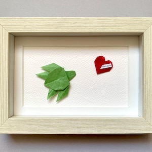 Può includere: Opera d'arte origami incorniciata con una tartaruga di carta verde e un cuore di carta rosso con la scritta "Alles Gute". L'opera è ambientata su uno sfondo bianco strutturato all'interno di una cornice in legno chiaro.