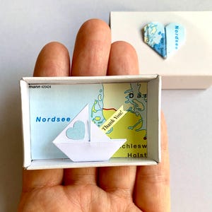 Könnte beinhalten: Eine kleine weiße Schachtel mit einem herzförmigen Kartendesign und eine Streichholzschachtel mit einem Papierschiff und einer Karte der Nordsee. Auf der Streichholzschachtel steht "Nordsee" und "Thank You!" auf einem gelben Etikett.
