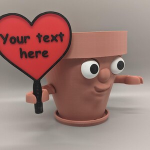 May include: A brown ceramic flower pot with a face, eyes, and a smile, holding a red heart-shaped sign that says "Your text here".