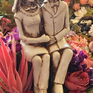 May include: A decorative figurine of a skeleton couple, seated and holding hands, surrounded by colorful flowers. The bride skeleton wears a veil and floral crown. The groom skeleton is in a suit. The base is a wood slice with moss.