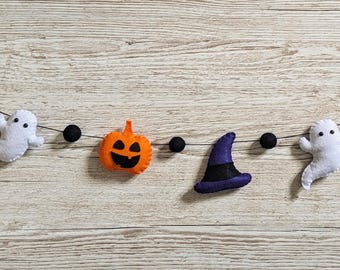 Puede incluir: Una guirnalda de Halloween con figuras de fieltro ensartadas en un cordel fino. La guirnalda incluye dos fantasmas blancos, dos calabazas naranjas con caras negras y dos sombreros de bruja morados, intercalados con pequeñas bolas de fieltro negras.