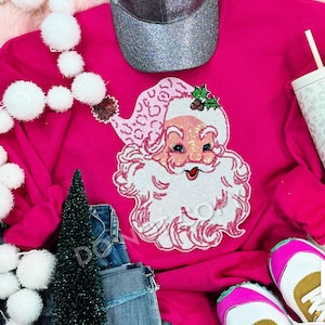 May include: A bright pink sweatshirt featuring a sequined Santa Claus design. The Santa has a white beard and a pink hat with a leopard print pattern. The image also includes a silver glitter baseball cap, jeans, and white sneakers with pink accents.