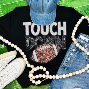 May include: A black long-sleeve shirt with sequined text reading TOUCH DOWN and a football graphic, a blue denim baseball cap, light blue denim shorts with silver buttons, white sneakers, and a long string of white and natural wood beads.
