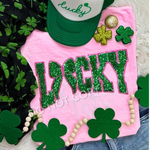May include: Pink t-shirt with the word "LUCKY" in green glitter letters. The letters are outlined in pink. The shirt is surrounded by green shamrocks and a white trucker hat with the word "lucky" embroidered on it.