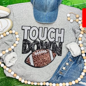 May include: A gray sweatshirt with the words "TOUCH DOWN" in large, sparkly letters and a football graphic. The image also includes a denim baseball cap, denim shorts, and white sneakers.