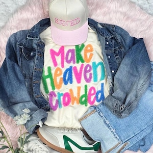 May include: A casual outfit featuring a cream-colored t-shirt with the phrase "Make Heaven Crowded" in colorful letters, paired with a denim jacket, light-wash jeans, a pink-billed cap with "SAVED" embroidered, and white sneakers.