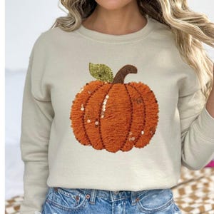 May include: Beige sweatshirt featuring an embroidered orange pumpkin with a green leaf and brown stem. The pumpkin design includes sequins. The sweatshirt is paired with blue denim shorts, creating a fall-themed outfit.