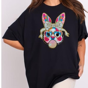 May include: Black oversized t-shirt featuring a colourful sequined bunny wearing sunglasses and blowing a bubble. The bunny has a pink nose and pink ears.