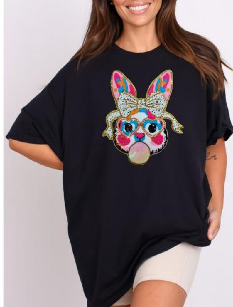 May include: Black oversized t-shirt featuring a colourful sequined bunny wearing sunglasses and blowing a bubble. The bunny has a pink nose and pink ears.