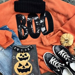 May include: An orange sweatshirt with the word "BOO" in black sequins, a black knit cap, a gray shirt with pumpkin appliques, blue jeans, and black sneakers with white soles. The theme is Halloween.