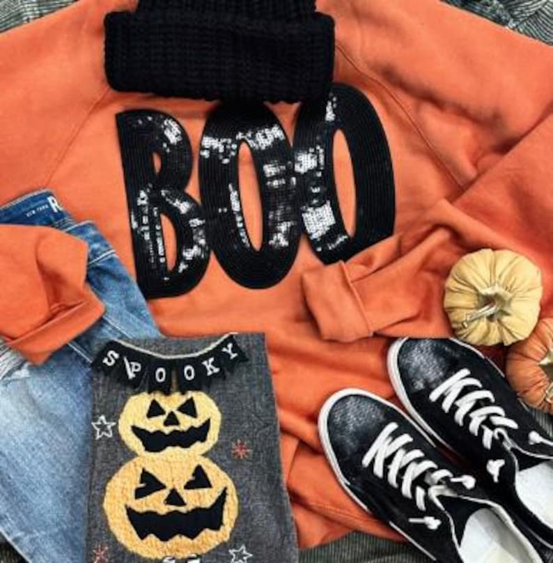May include: An orange sweatshirt with the word "BOO" in black sequins, a black knit cap, a gray shirt with pumpkin appliques, blue jeans, and black sneakers with white soles. The theme is Halloween.