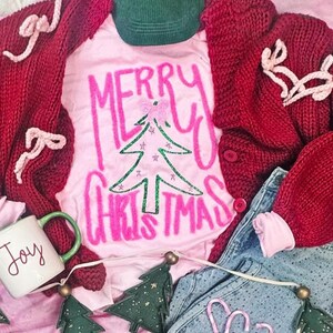 May include: A festive holiday scene featuring a pink t-shirt with "MERRY CHRISTMAS" and a Christmas tree graphic. A red cardigan, green corduroy hat, denim jeans, and a mug that says "Joy" are also visible. Decorative Christmas tree ornaments complete the arrangement.