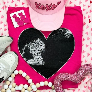 May include: A pink t-shirt with a large black sequined heart design. A pink baseball cap with "XOXO" text sits above the shirt. The image also includes pink heart earrings, a beaded necklace, and gray sneakers.