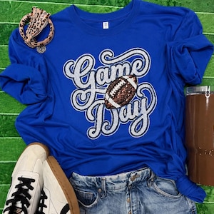 May include: A royal blue long-sleeve t-shirt featuring the silver glitter text "Game Day" with a brown and white football graphic replacing the letter "a". The shirt is paired with blue denim jeans, white sneakers, and a leopard print hair tie.
