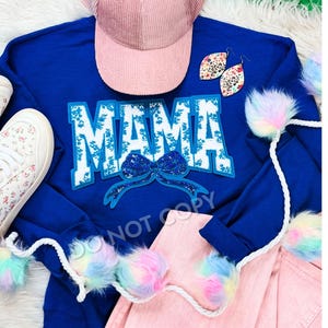 May include: A royal blue sweatshirt with the word "MAMA" in white letters with a blue and silver bow. The sweatshirt is laid out on a pink surface with white rope and colourful pom-poms.