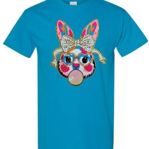 May include: A turquoise blue t-shirt featuring a colourful graphic of a cartoon bunny wearing heart-shaped sunglasses and a bow. The bunny is blowing a bubble.