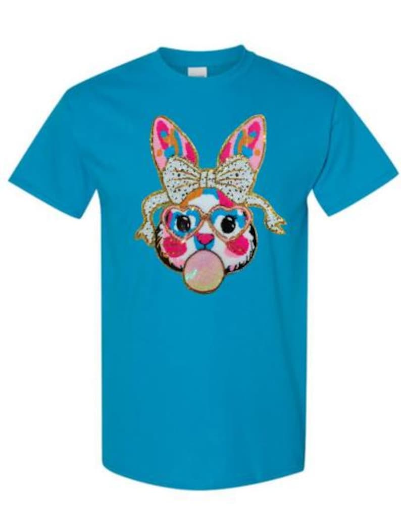 May include: A turquoise blue t-shirt featuring a colourful graphic of a cartoon bunny wearing heart-shaped sunglasses and a bow. The bunny is blowing a bubble.