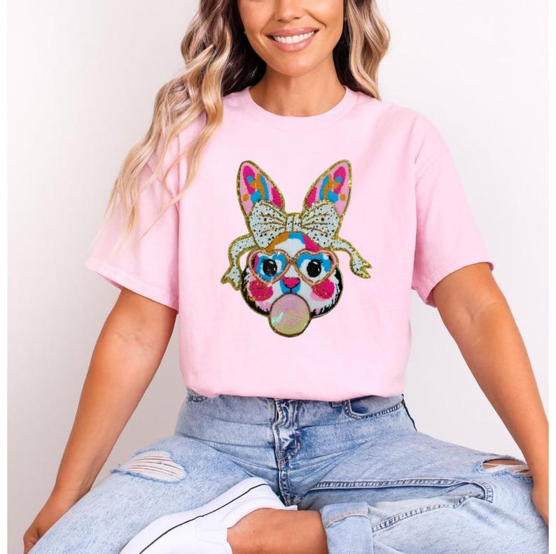 May include: A pink t-shirt featuring a colourful sequined bunny wearing sunglasses and blowing a bubble. The bunny has a pink nose and white whiskers.
