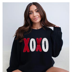 May include: Black sweatshirt with the letters XOXO in red and white. The letters have a fuzzy texture. The sweatshirt is a crew neck style and has long sleeves. The background is a plain white.