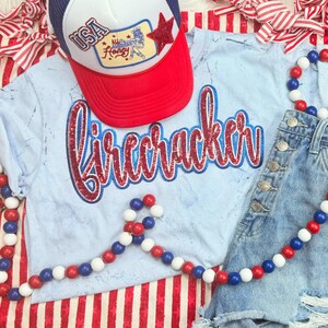 May include: A light blue t-shirt with the word "Firecracker" in red and blue glitter lettering, paired with denim shorts and a red, white, and blue trucker hat. A red, white, and blue beaded garland adds a festive touch.