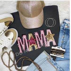 Può includere: Una felpa nera con la parola "MAMA" in paillettes rosa e dorate con una stella rosa e dorata sulla "A". La felpa è appoggiata su un tavolo con un cappello da camionista con paillettes dorate, un paio di scarpe da ginnastica bianche con dettagli dorati, un paio di orecchini a cerchio dorati, una collana di perline di legno e un paio di jeans blu con bottoni dorati.