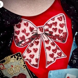 May include: A red velvet shirt with a large pink and red sequined bow. The bow is made of hearts and is a statement piece.