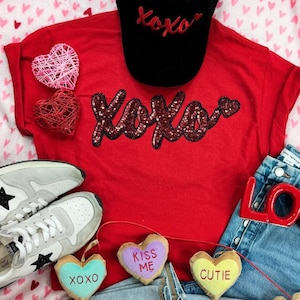 May include: A red t-shirt with the word "XOXO" in sequined letters, paired with a black cap with "Xoxo" embroidered on it. The image also includes a pair of gray sneakers with black stars, denim shorts, and heart-shaped decorations.