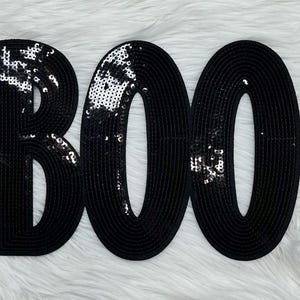 May include: Black sequined "BOO" letters on a white fluffy surface. The letters are outlined in black and filled with black sequins. The sequins catch the light, creating a sparkling effect. The design is perfect for Halloween decorations.