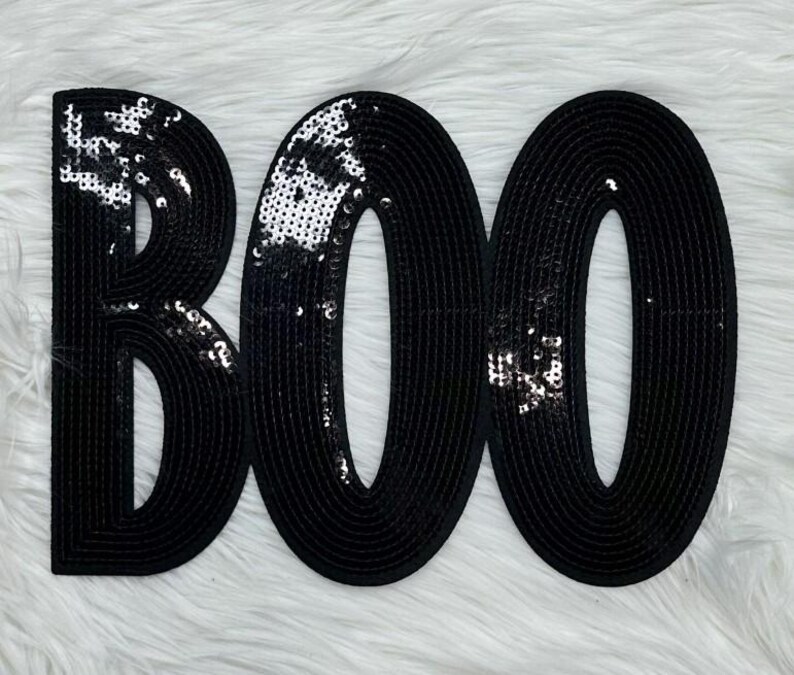 May include: Black sequined "BOO" letters on a white fluffy surface. The letters are outlined in black and filled with black sequins. The sequins catch the light, creating a sparkling effect. The design is perfect for Halloween decorations.