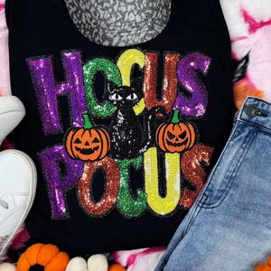 Può includere: Canotta nera con la scritta "Hocus Pocus" in lettere paillettate. Il design include un gatto nero e due zucche. La maglietta è abbinata a jeans blu e scarpe da ginnastica bianche, suggerendo un tema di Halloween.