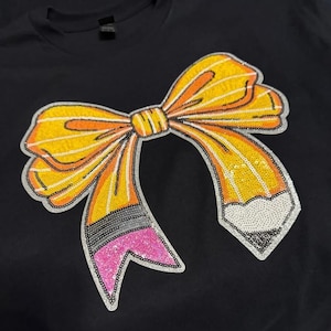 May include: A black t-shirt featuring a large, decorative bow graphic made of sequins. The bow is designed to look like a yellow pencil with orange stripes, a grey metal ferrule, a pink eraser, and a white pencil tip, all outlined in silver.