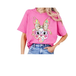 May include: Pink t-shirt featuring a colourful bunny wearing heart-shaped sunglasses and a polka dot bow. The bunny is holding a bubble.