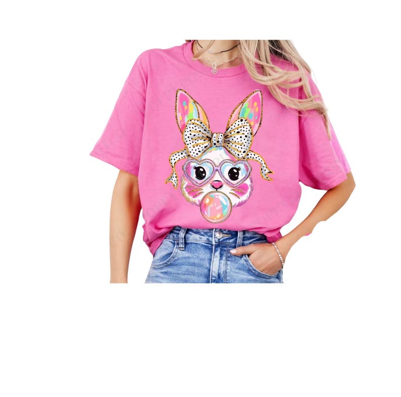 May include: Pink t-shirt featuring a colourful bunny wearing heart-shaped sunglasses and a polka dot bow. The bunny is holding a bubble.