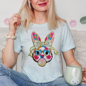 May include: A light blue t-shirt featuring a colourful sequined bunny wearing sunglasses and blowing a bubble. The bunny has a pink nose, blue eyes, and a gold bow. The text "Not Too" is visible on the shirt.