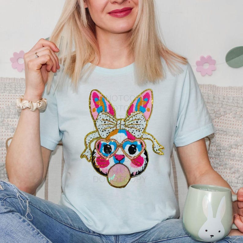 May include: A light blue t-shirt featuring a colourful sequined bunny wearing sunglasses and blowing a bubble. The bunny has a pink nose, blue eyes, and a gold bow. The text "Not Too" is visible on the shirt.