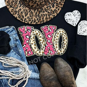 May include: Black sweatshirt with a pink and gold "XOXO" graphic featuring a leopard print design. The sweatshirt is paired with blue jeans, a brown hat, and white decorative hearts.